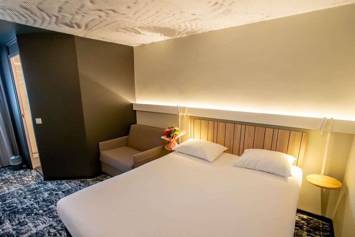 A hotel room with a large bed, a sofa, a small table with a vase of flowers, and a nightstand with a lamp.