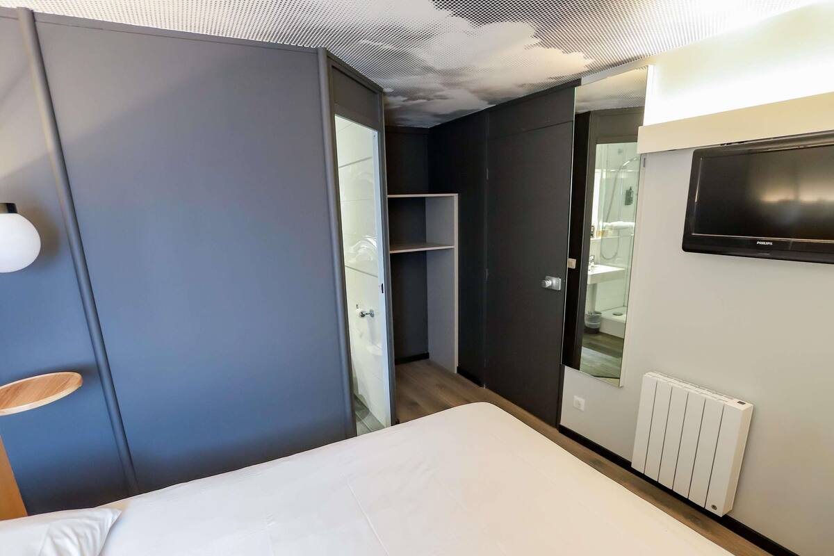 A modern hotel room with a bed, a TV mounted on the wall, a wardrobe, and a bathroom visible through an open door.