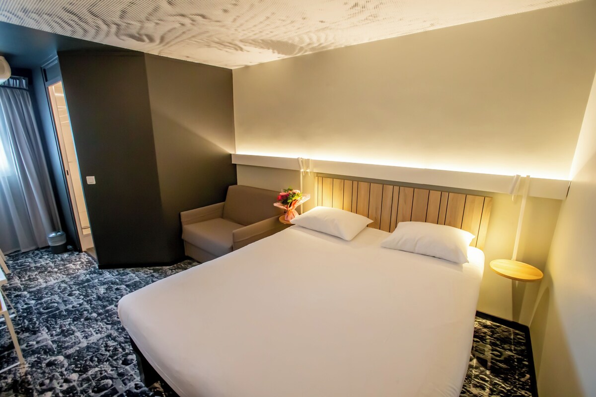 A hotel room with a large bed, a small sofa, a bedside table with a lamp, and a wooden headboard.