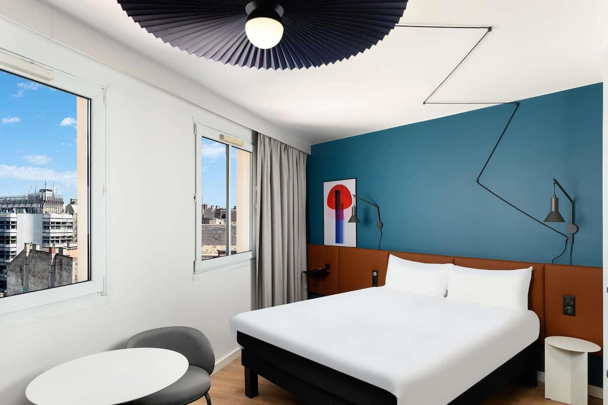 A hotel room with a bed, a chair, a small table, a city view, and a ceiling fan.