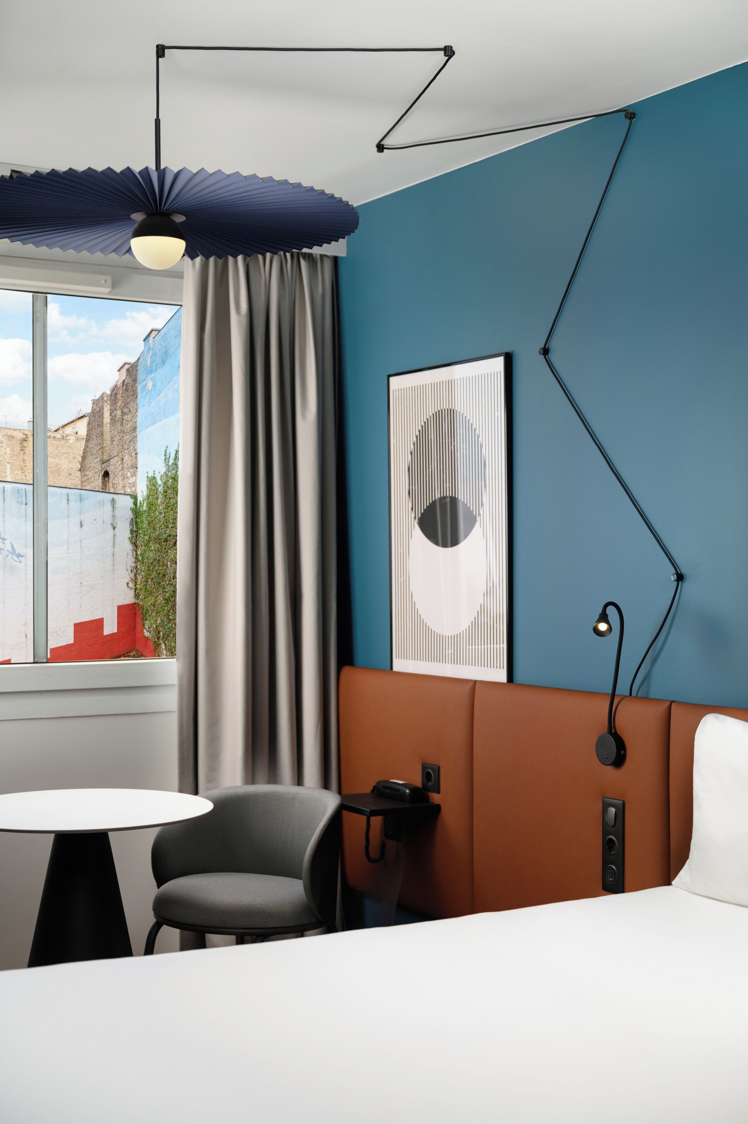 Una camera d'albergo con un letto, una sedia, una scrivania, una lampada, una finestra con tende e un'opera d'arte appesa al muro.