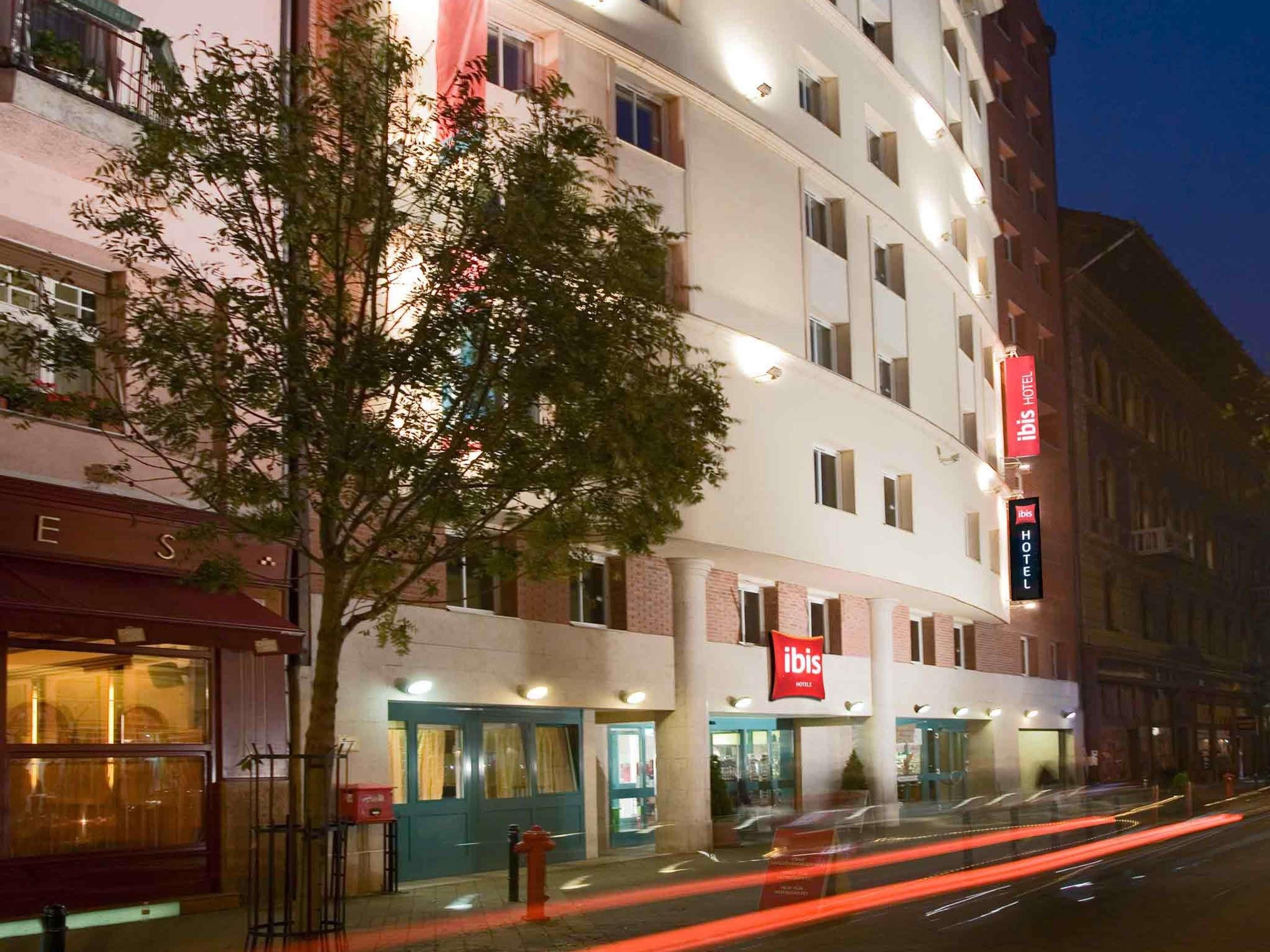 A multi-story hotel with a prominent 'ibis HOTEL' sign, a tree in front, and a street with light trails.