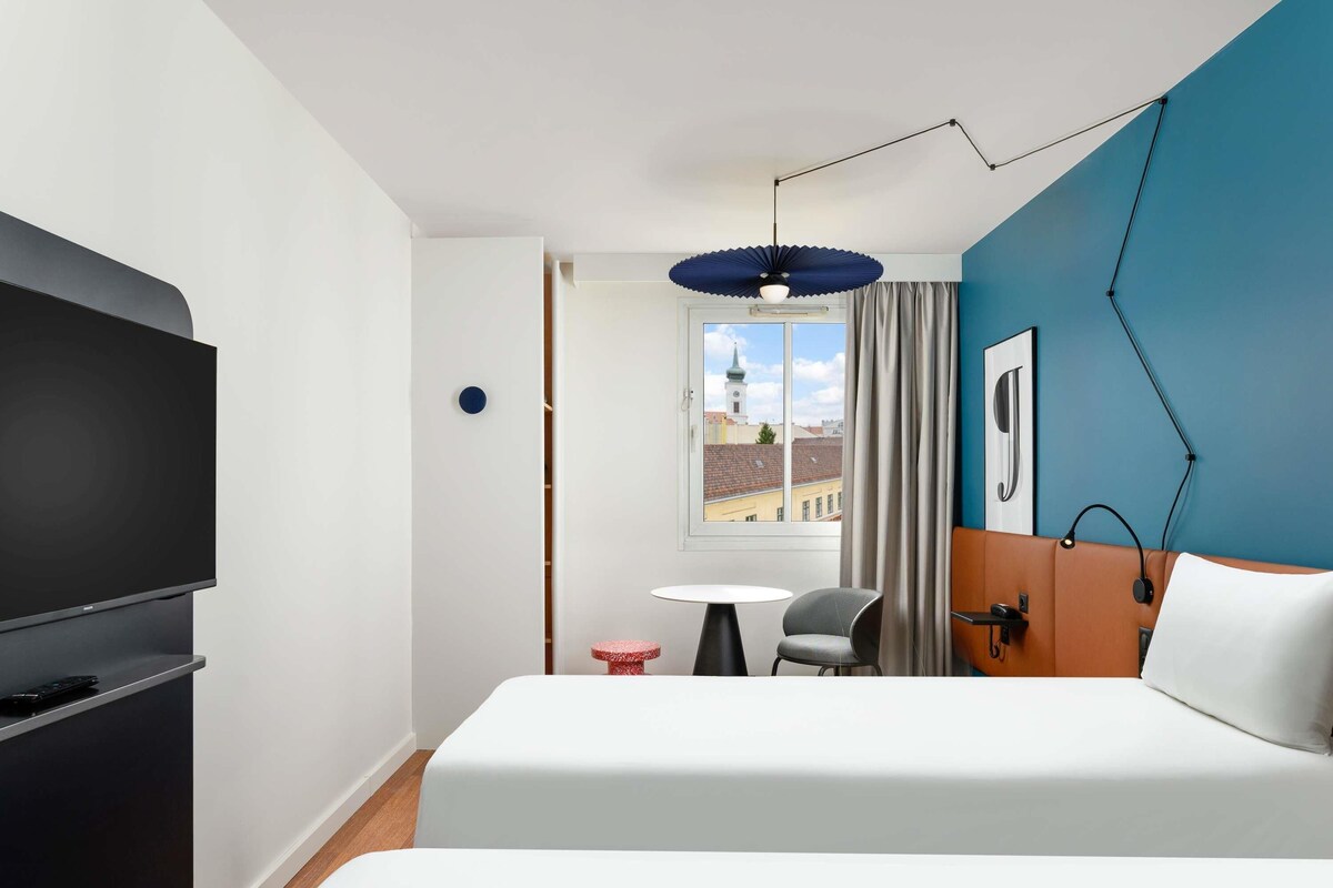 Une chambre d’hôtel comprenant un lit, une télévision, un bureau, une chaise, une fenêtre donnant sur des immeubles et un mur d’accent bleu.