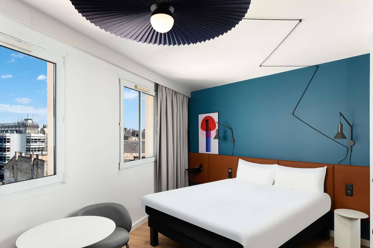 A modern hotel room with a large bed, a small table, a chair, a city view through a window, and a unique ceiling light fixture.