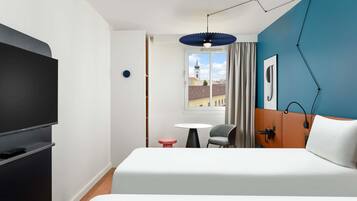 Habitación de hotel con cama, televisor, escritorio, silla y ventana que ofrece vistas a edificios, además de una pared decorada en azul.