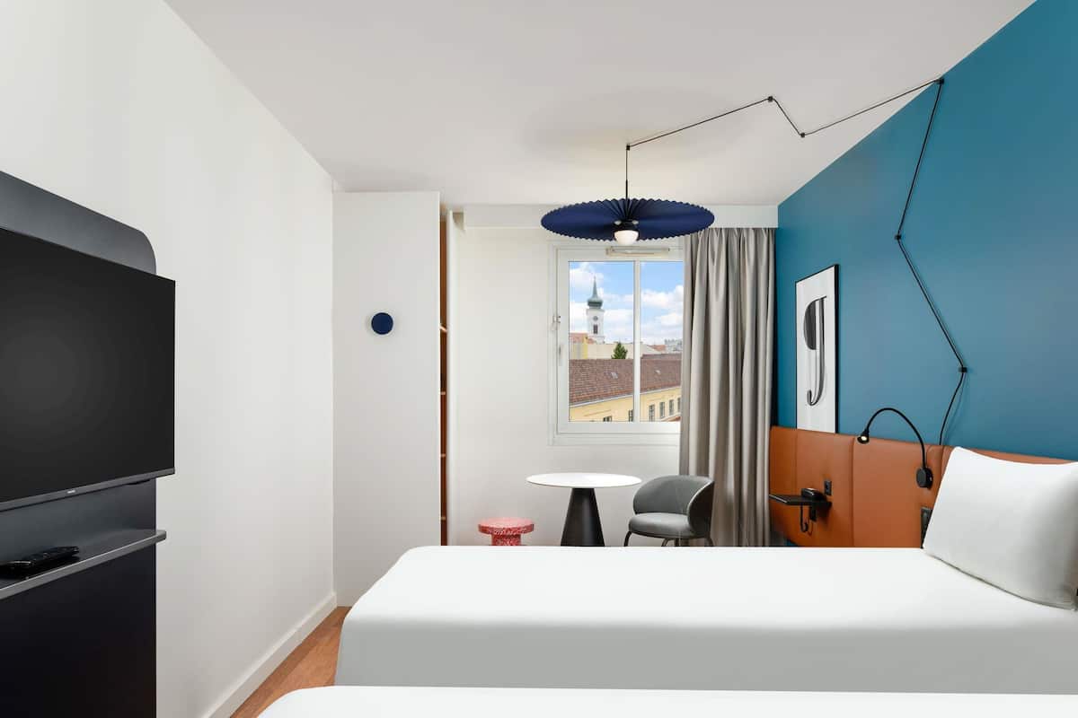 A hotel room with a bed, a TV, a desk, a chair, a window with a view of buildings, and a blue accent wall.