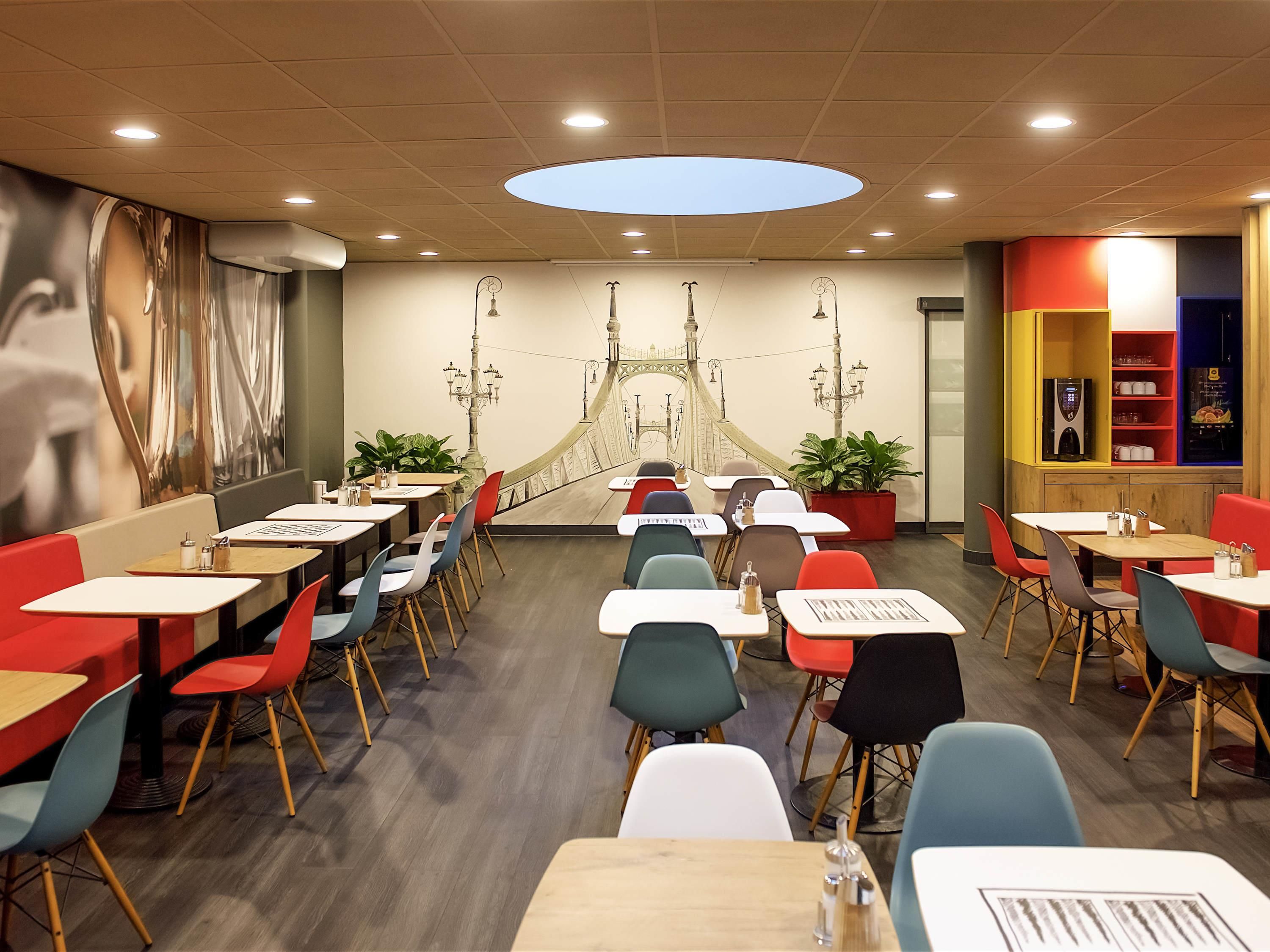 Un restaurante con un mural de un puente, varias mesas y sillas, y un diseño moderno en el techo.