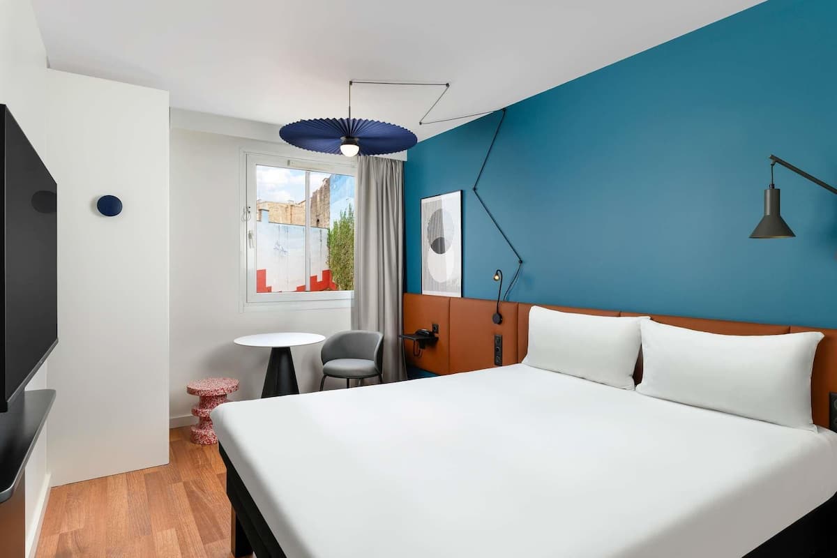 A hotel room with a bed, a desk, a chair, a small table, a TV, and a window with curtains.
