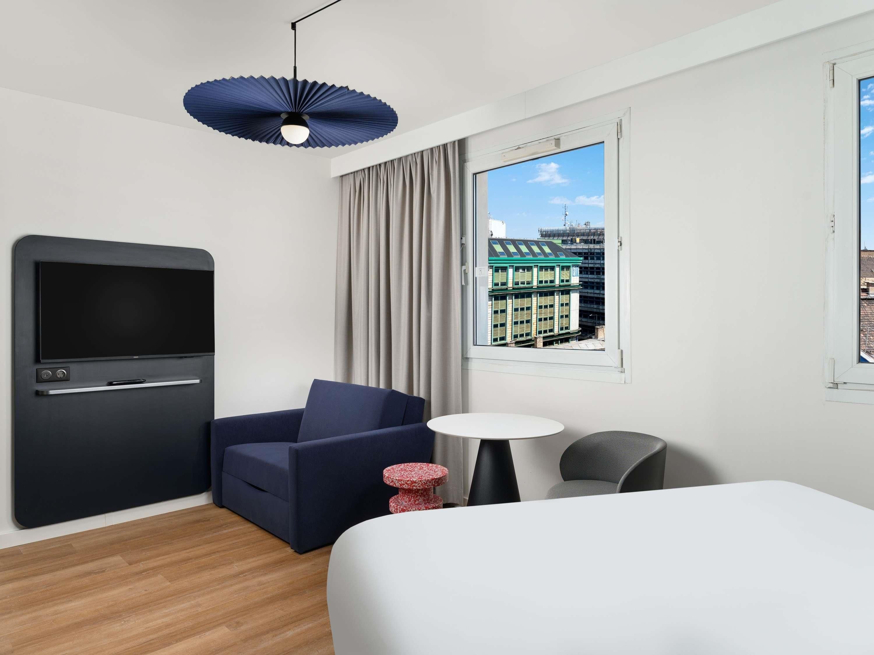 Ett hotellrum med en säng, en tv, en soffa, ett litet bord, en lampa och utsikt över byggnaderna utanför.