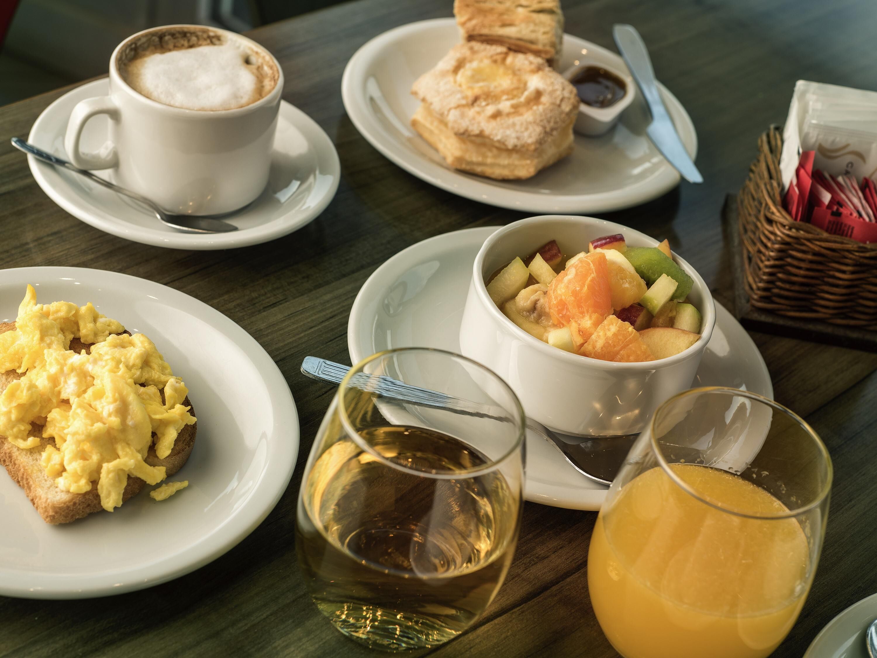A breakfast setting with scrambled eggs, a cup of coffee, pastries, a fruit bowl, and glasses of wine and orange juice.