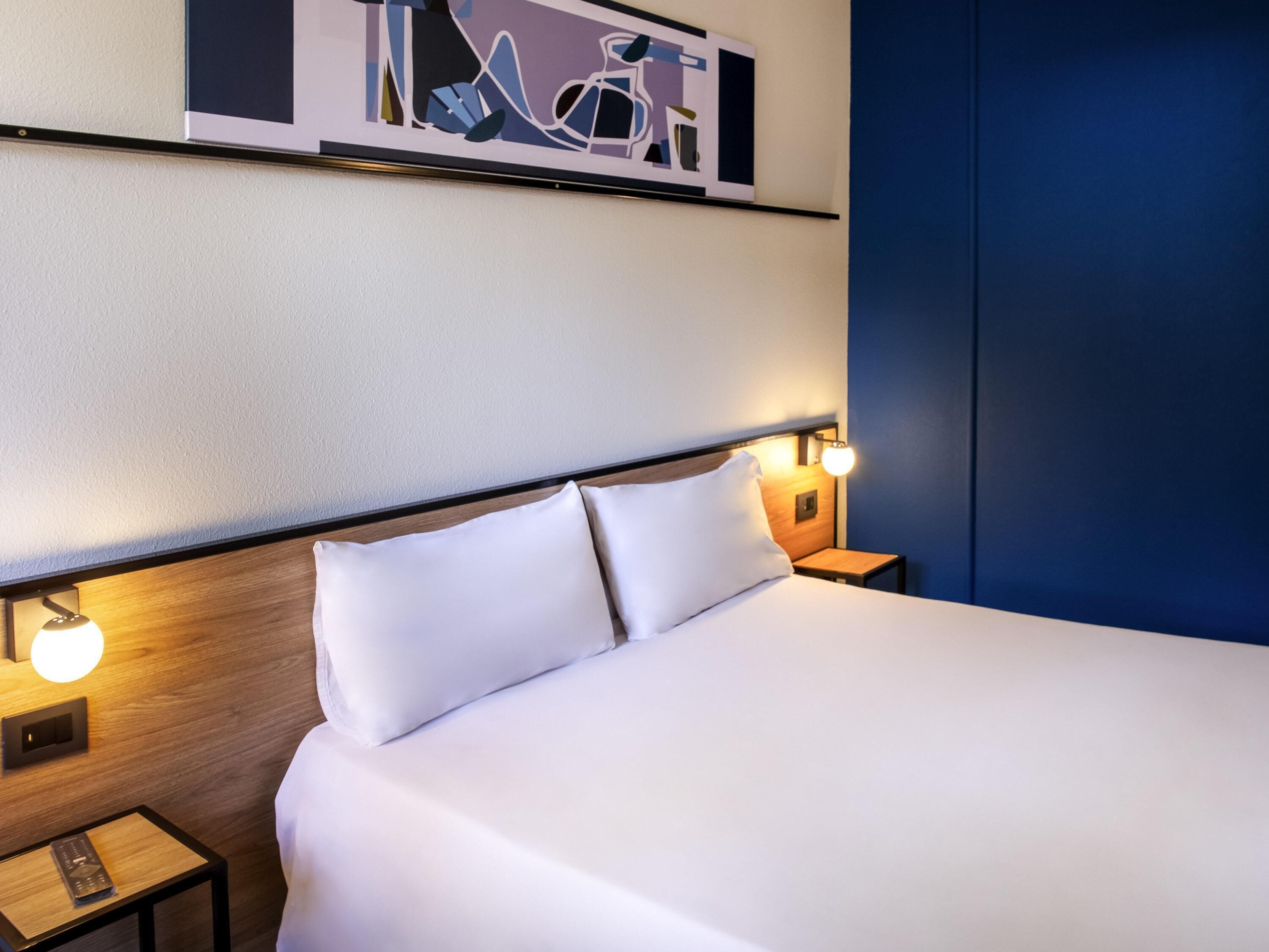 Cama bem arrumada com lençóis brancos, cabeceira com interruptor de luz e duas luminárias de mesa.
