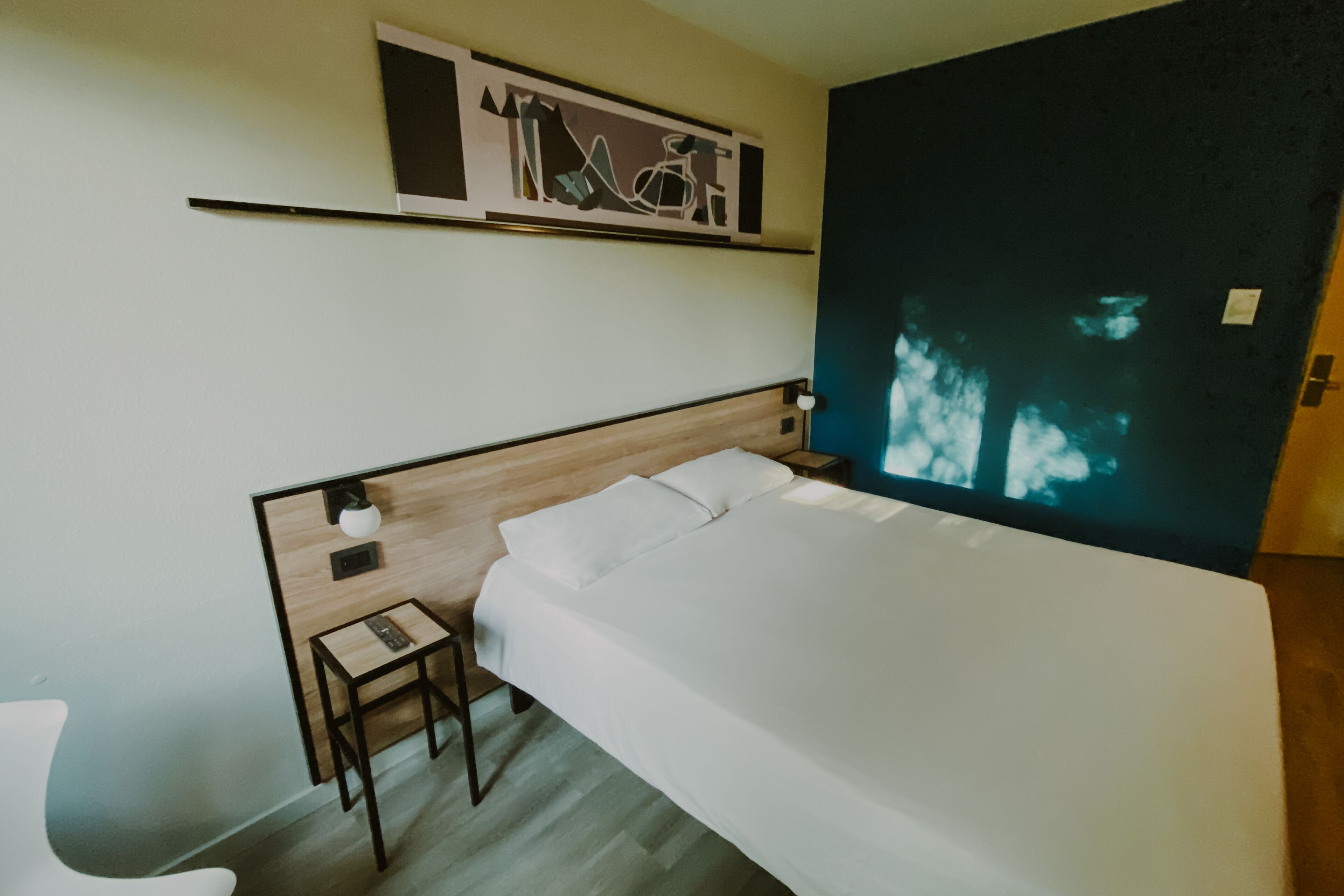 A modern hotel room with a wooden headboard, a bed with white linens, a small bedside table with a lamp, and a wall-mounted artwork above the bed.