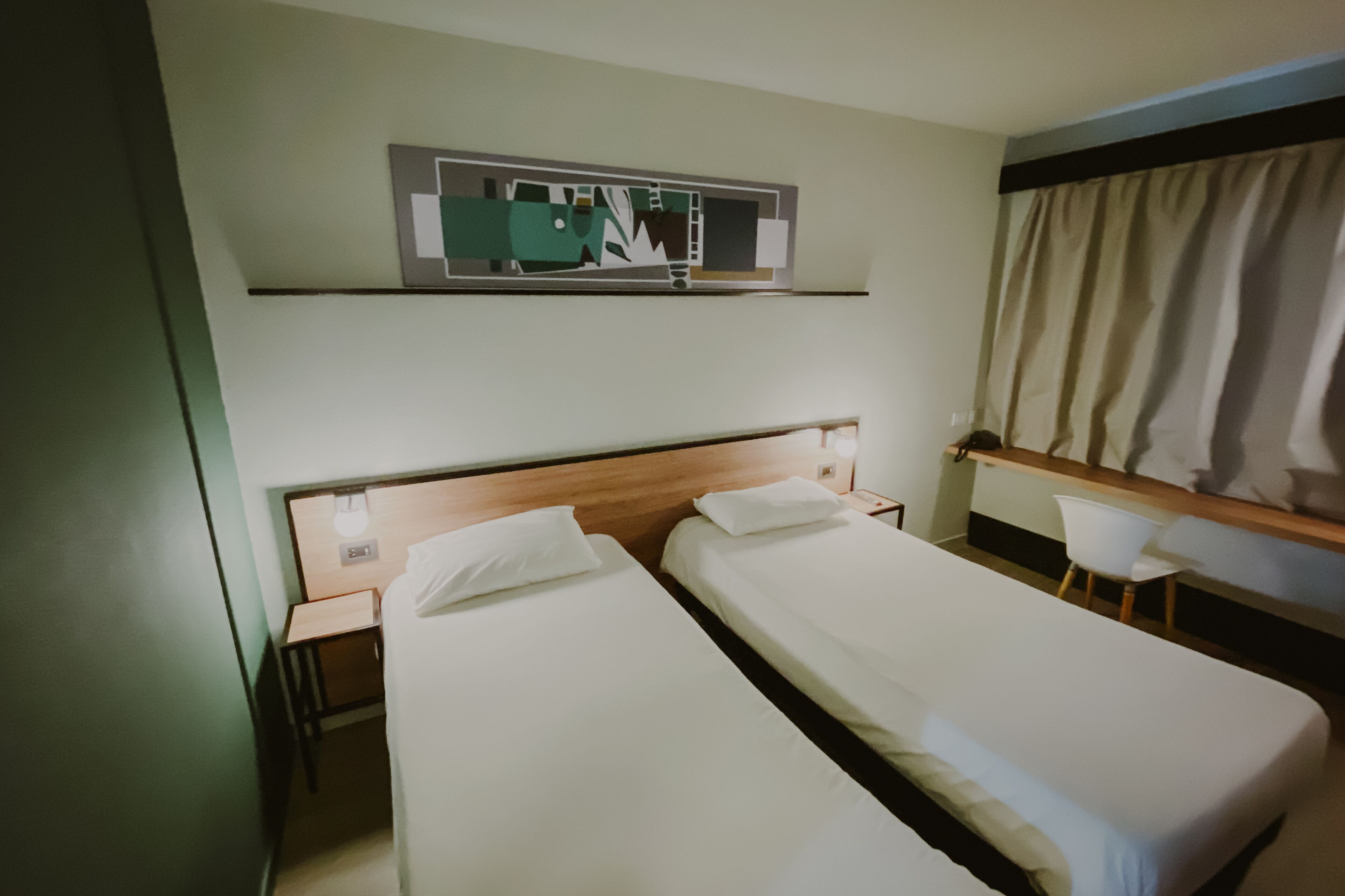 A hotel room with two single beds, a desk, a chair, a mirror, and a window with curtains.