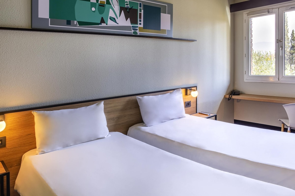 Ein Hotelzimmer mit zwei Einzelbetten, einem Schreibtisch, einem Stuhl, einem Wandbild und einem Fenster mit Blick auf Bäume.