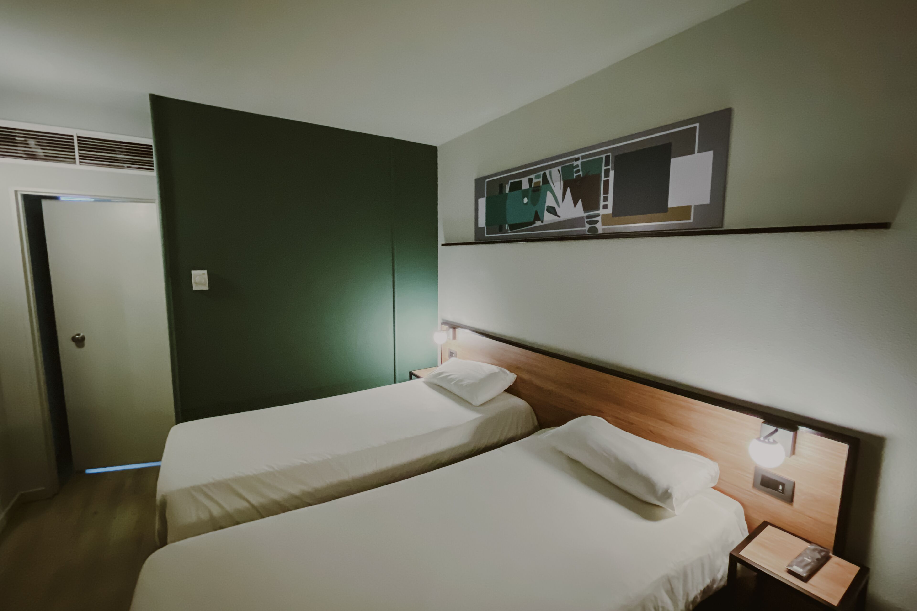 A hotel room with two single beds, a wooden headboard, a nightstand with a lamp, and a wall-mounted artwork.