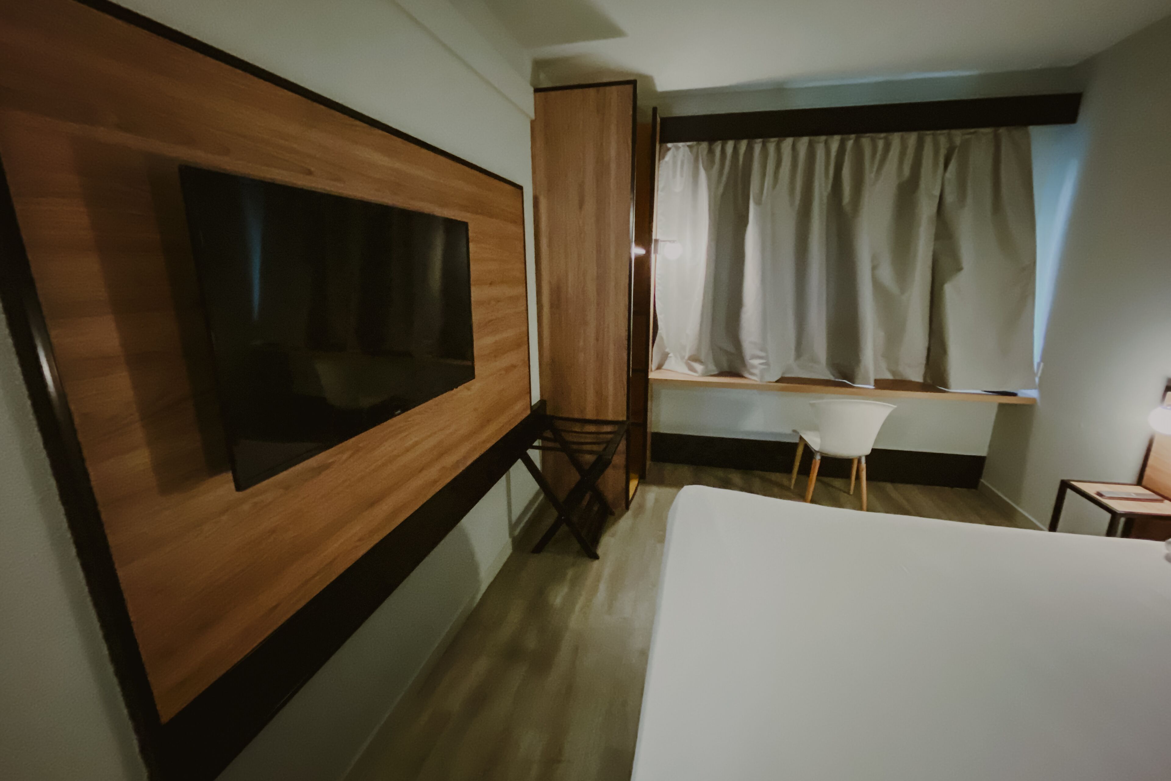 Habitación de hotel con una cama grande, un televisor montado en la pared, una mesita, una silla y un ventanal con cortinas.