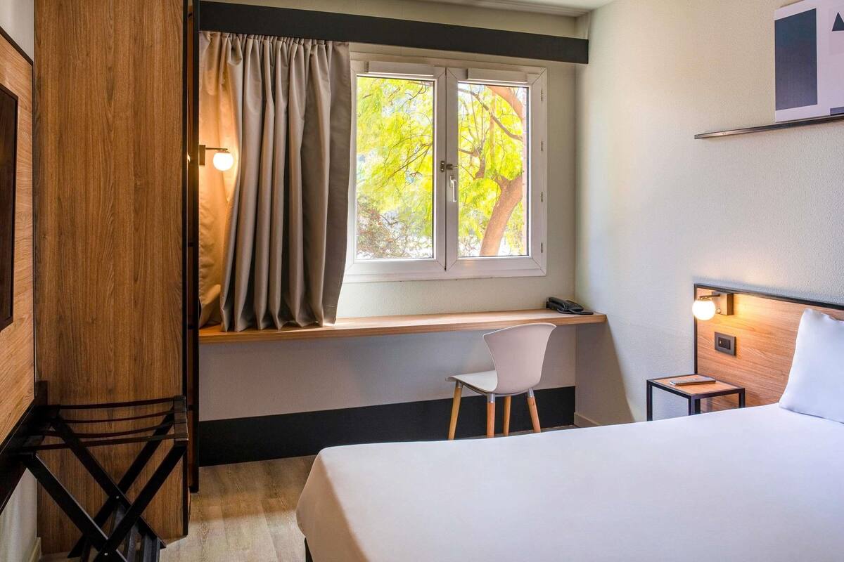 Ein Hotelzimmer mit einem Bett, einem Schreibtisch mit Stuhl, einem Fenster mit Vorhängen und einem Nachttisch mit Lampe.