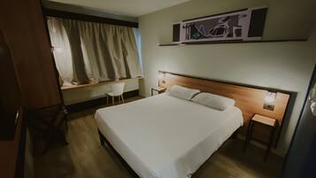 A hotel room with a large bed, a desk, a chair, a window with curtains, and a wall-mounted artwork.