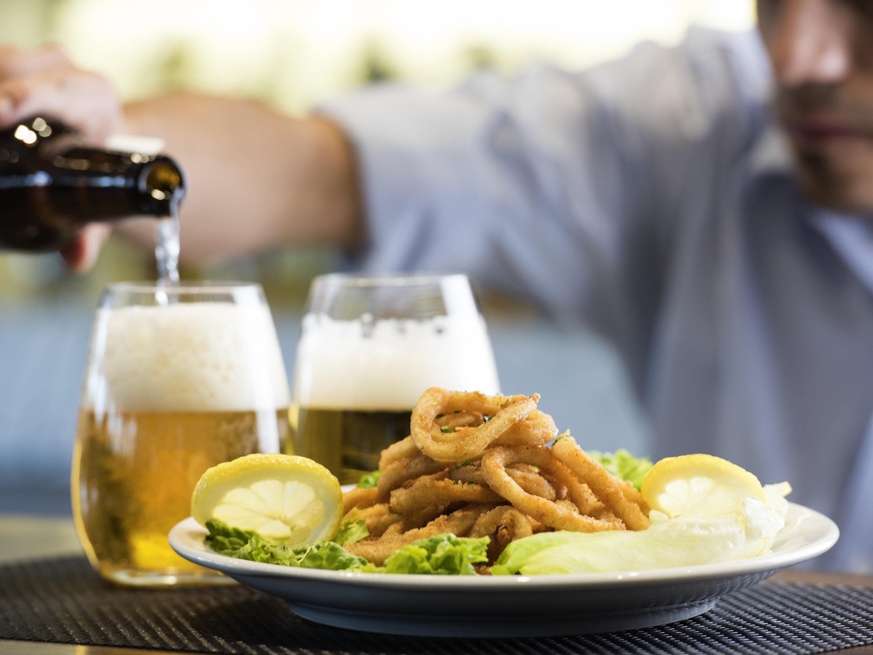 A plate of fried calamari with lemon wedges and lettuce, two glasses of beer, and a person pouring a drink.
