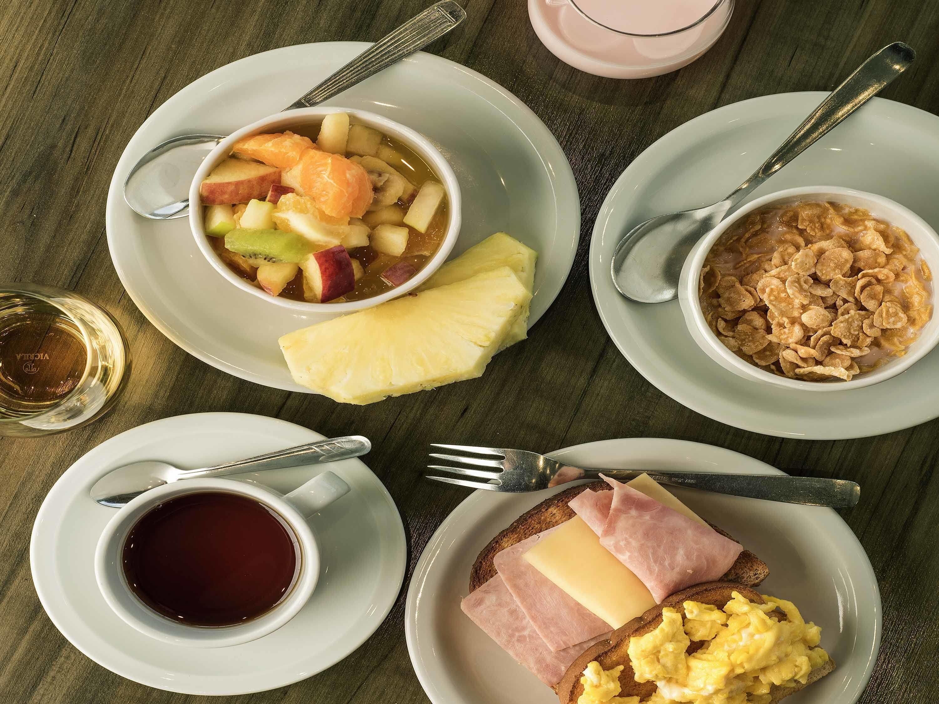 A breakfast setting with a bowl of fruit, a cup of coffee, cereal, toast with cheese and ham, and a glass of juice.