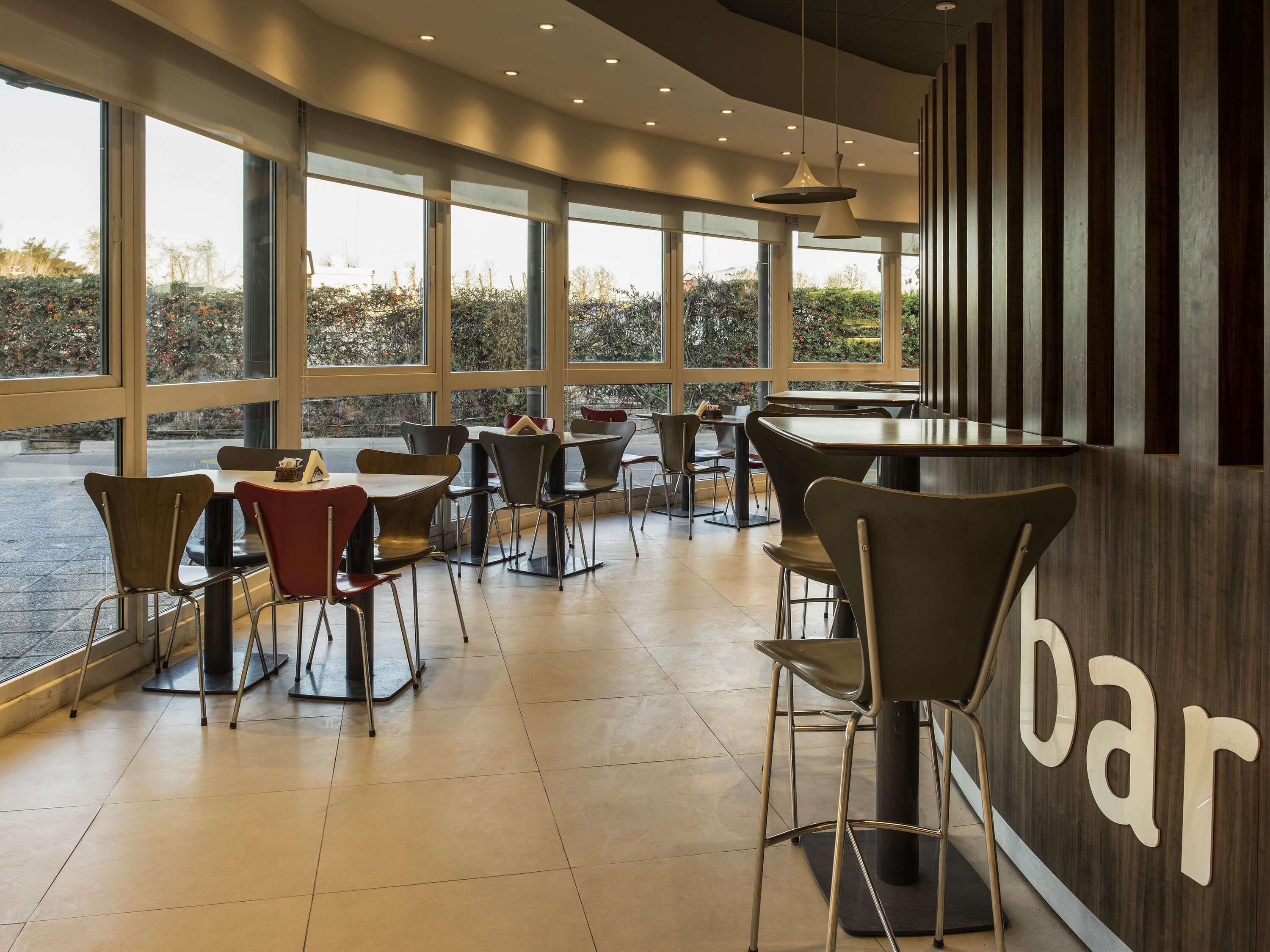 A modern bar with large windows, metal chairs, and a tiled floor.