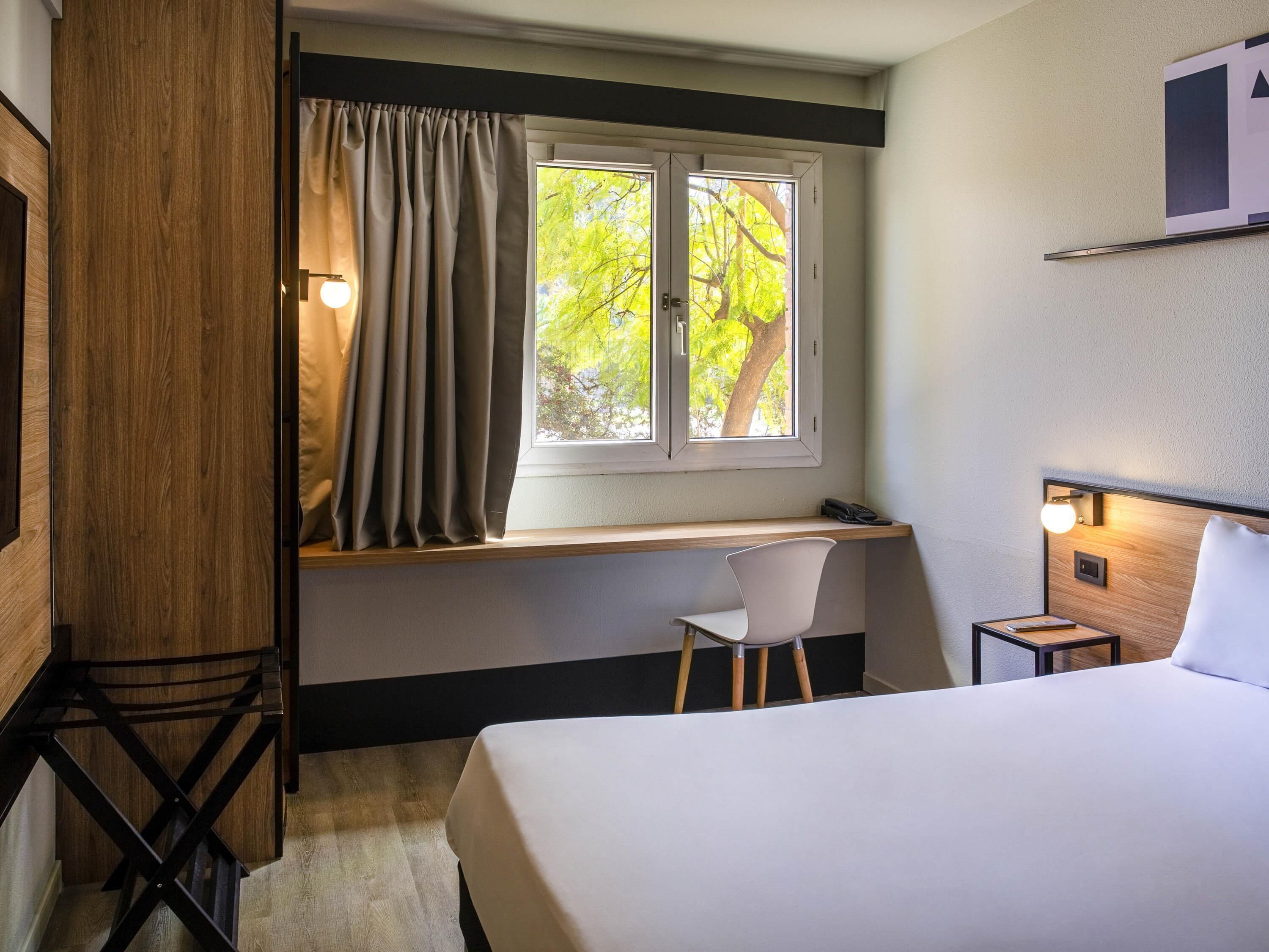 A hotel room with a bed, a desk, a chair, a nightstand, a window with curtains, and a wall-mounted shelf.