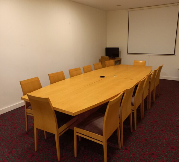 Un salón de conferencias con una larga mesa de madera y sillas a juego, una pantalla de proyector y piso alfombrado.