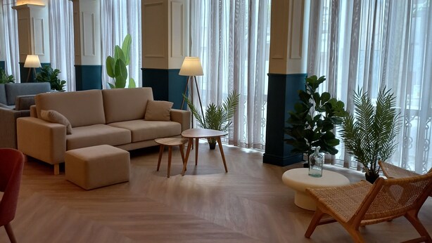 Amplia sala de estar con un conjunto de sofás, una mesa redonda de centro y una silla de mimbre. Se observan ventanas grandes con cortinas traslúcidas, una lámpara de pie y varias plantas en macetas.