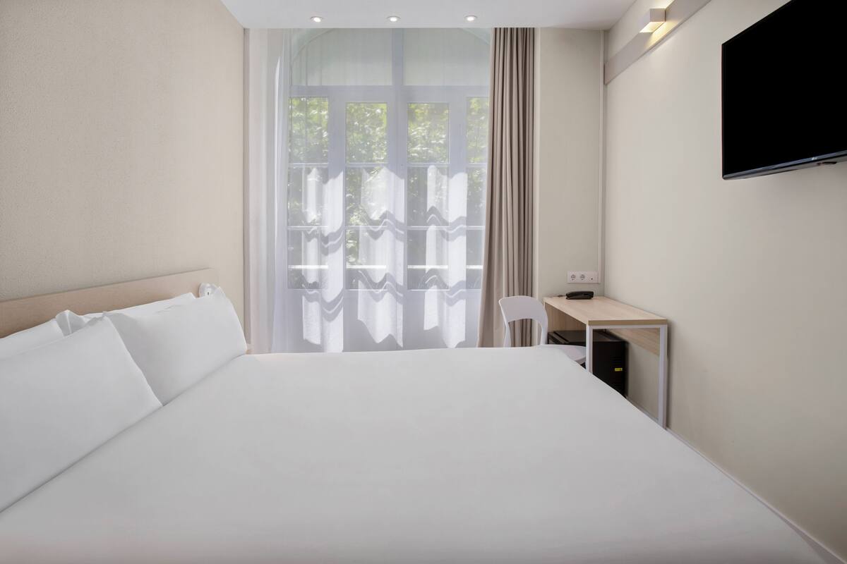 Una habitación de hotel con una cama grande, un escritorio, una silla, un televisor y una ventana con cortinas.