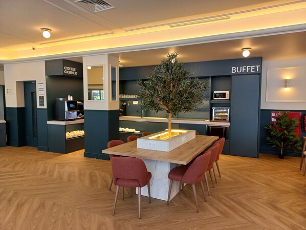 Een moderne hotel lobby met een centrale houten tafel, rode stoelen, een buffetgedeelte en een koffiehoek.