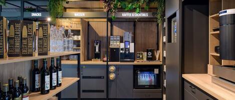 Zona de bar con estantes de bebidas embotelladas, una máquina de café y un letrero que anuncia aperitivos y agua.