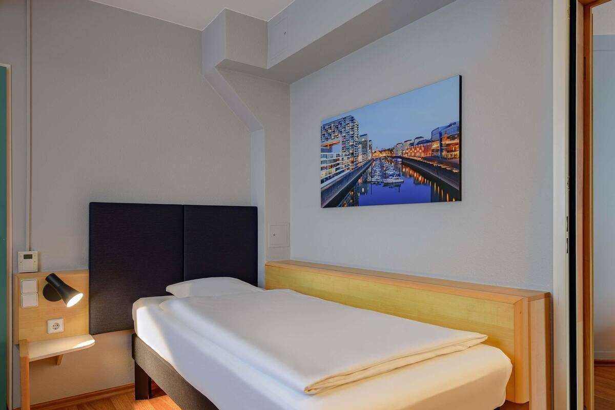 A hotel room with a bed, bedside lamp, and a cityscape painting on the wall.