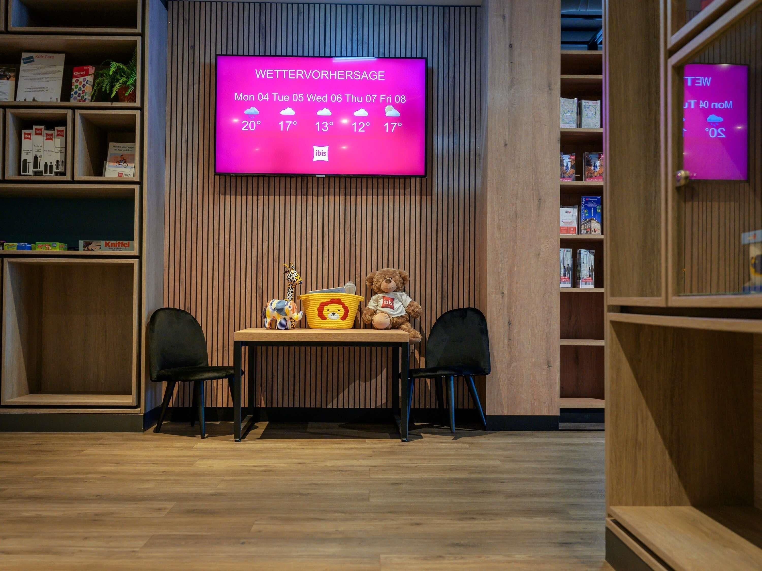 A room with a wooden floor, a table with chairs, a teddy bear, a basket, and a television displaying a weather forecast.