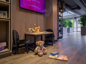 A modern interior with a wooden wall, a television displaying a message, a table with chairs, and a teddy bear on the floor. - ibis Köln Frechen (Frechen)