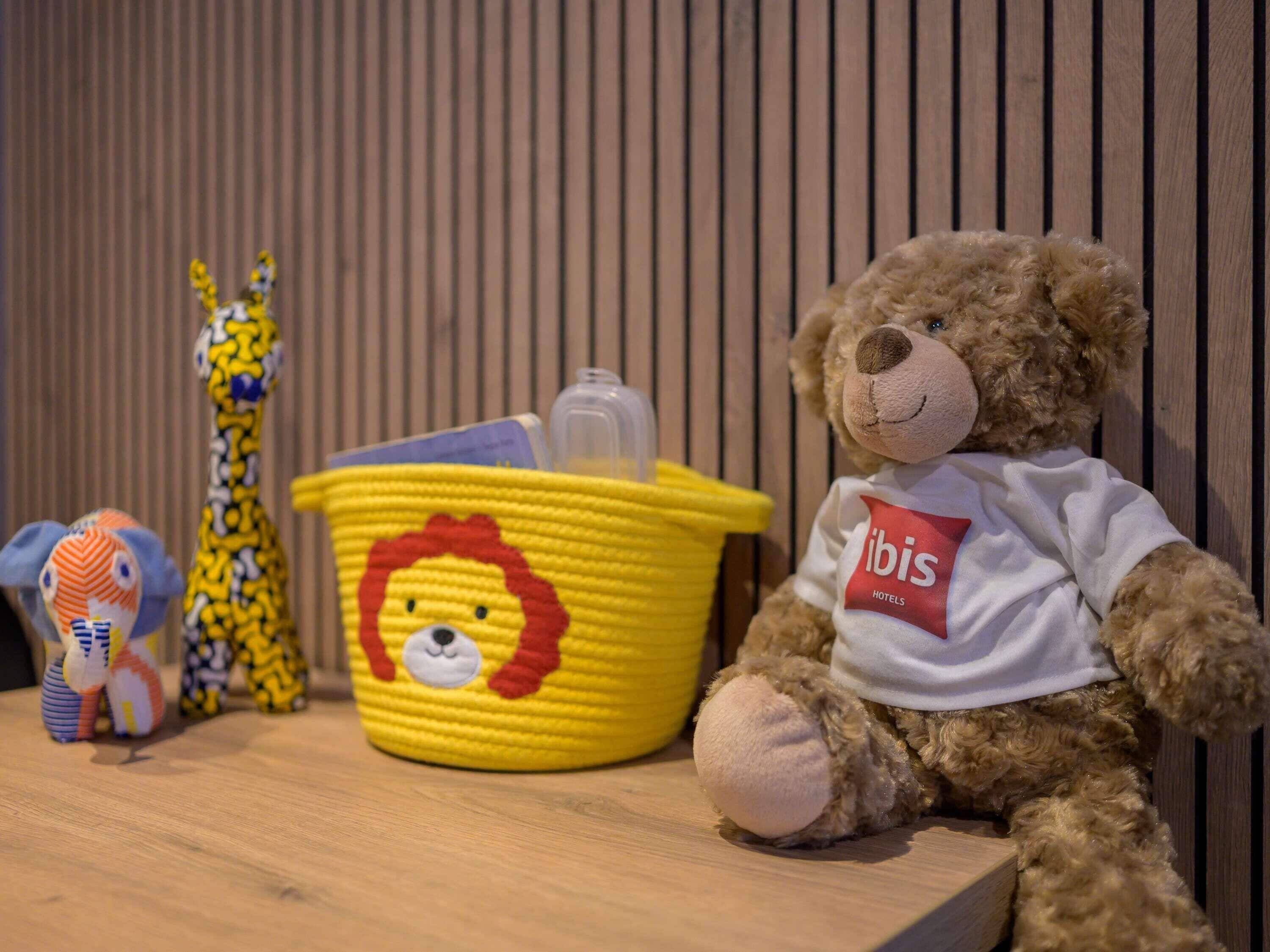 Un oso de peluche con una camiseta de 'libis hotels', una cesta amarilla con un diseño de león y un juguete de jirafa sobre una superficie de madera.