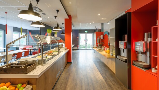 A modern hotel lobby with a long reception desk, a coffee machine, and a buffet area with fruits and vegetables.