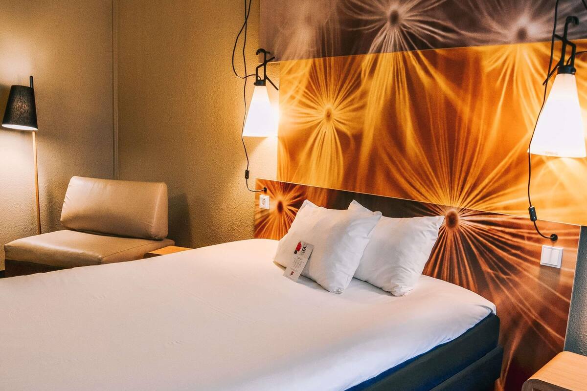 A hotel room with a bed, two bedside lamps, a chair, and a wall with a radiant design.