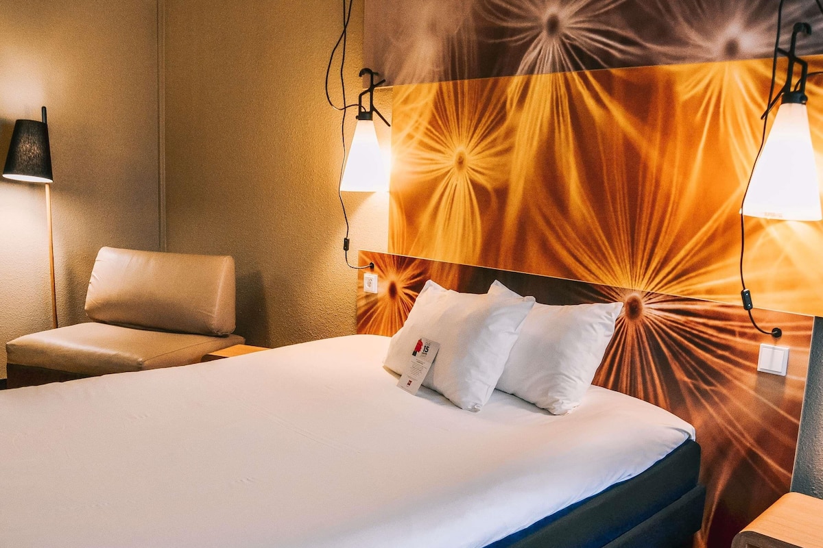 Quarto de hotel com uma cama, duas luminárias laterais, uma cadeira e parede com design radiante.