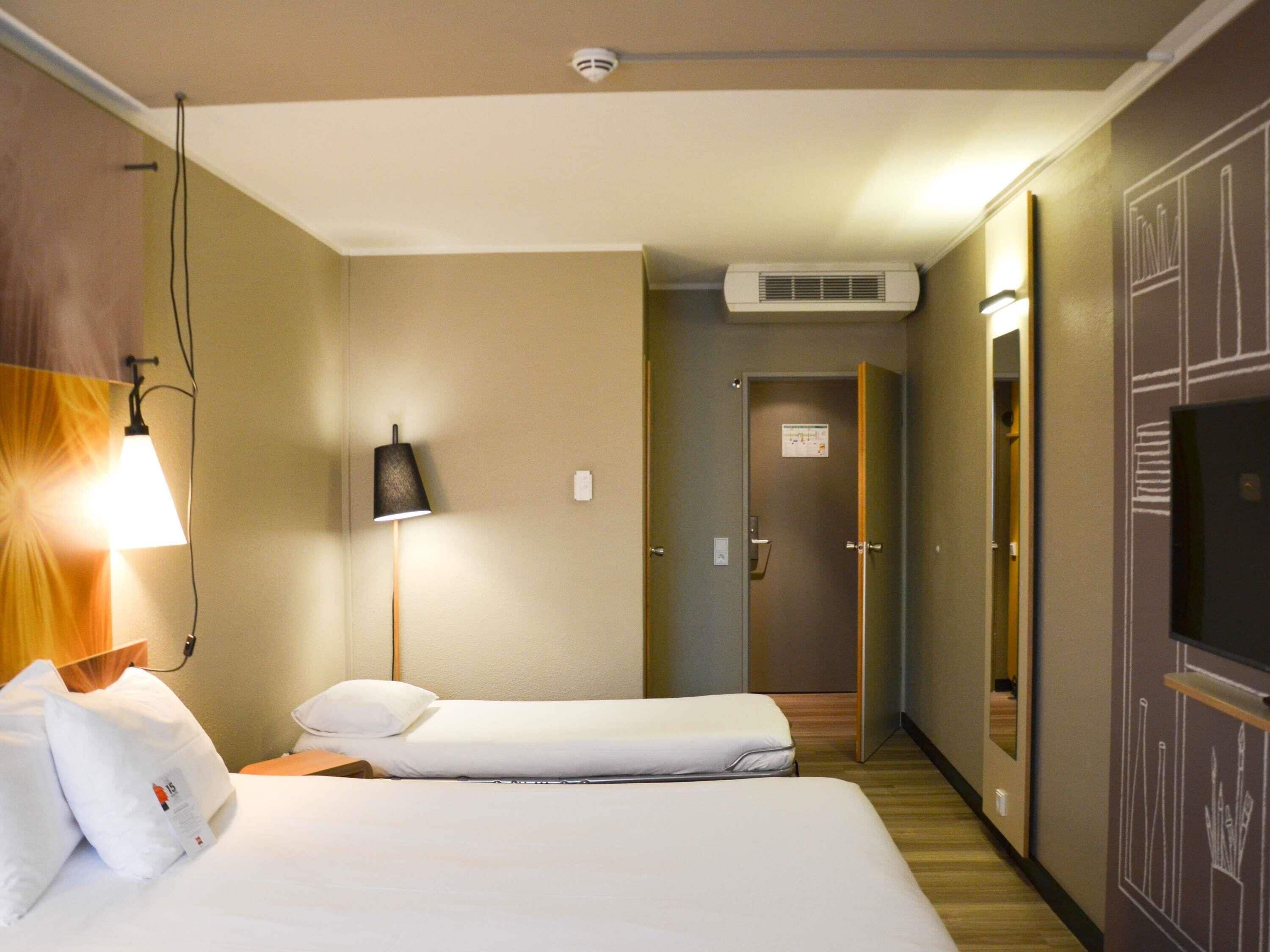A hotel room with a bed, bedside lamp, air conditioning unit, and a mirror.