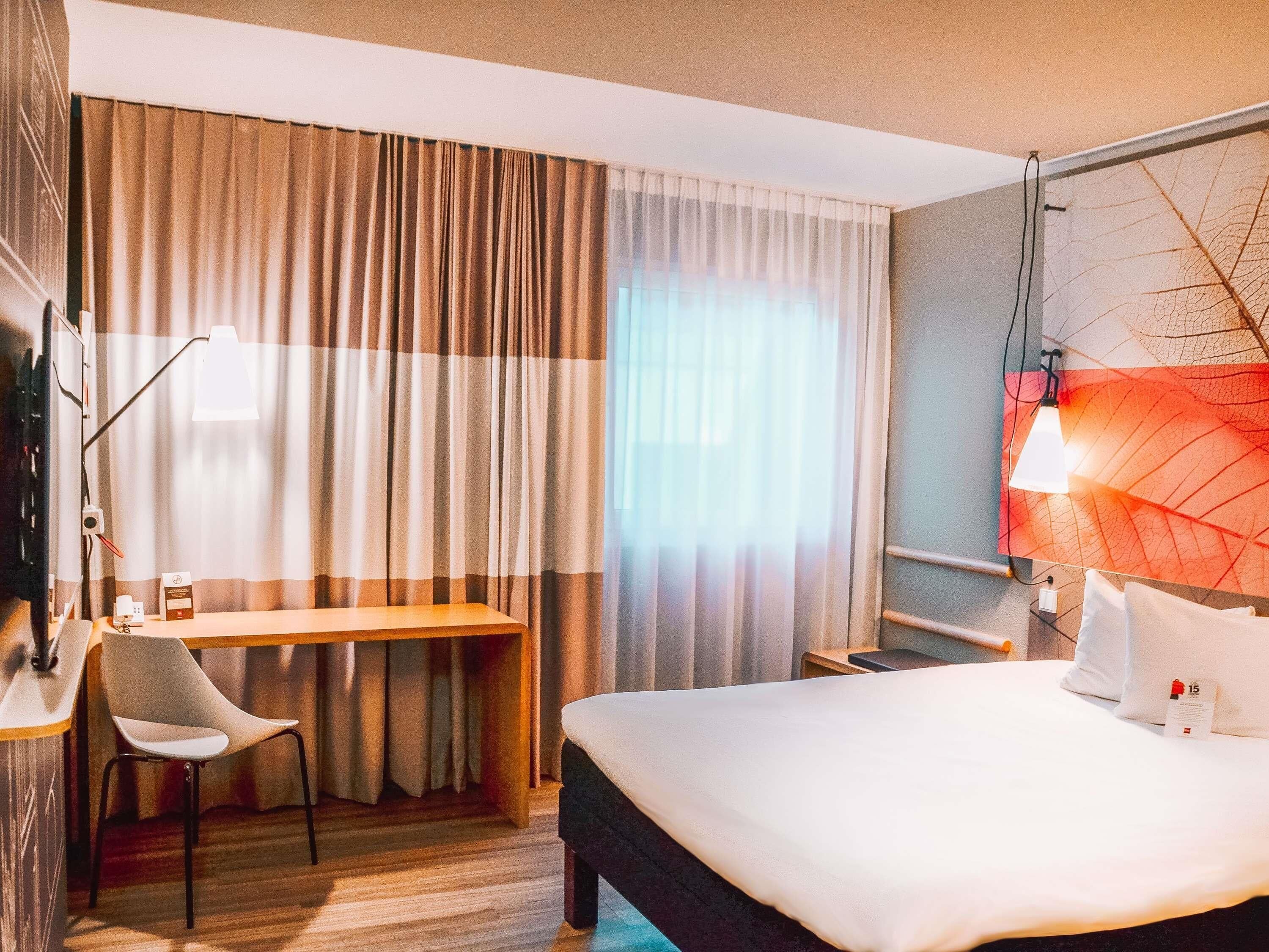 Habitación de hotel con cama, escritorio, silla, televisor y lámpara de pared.