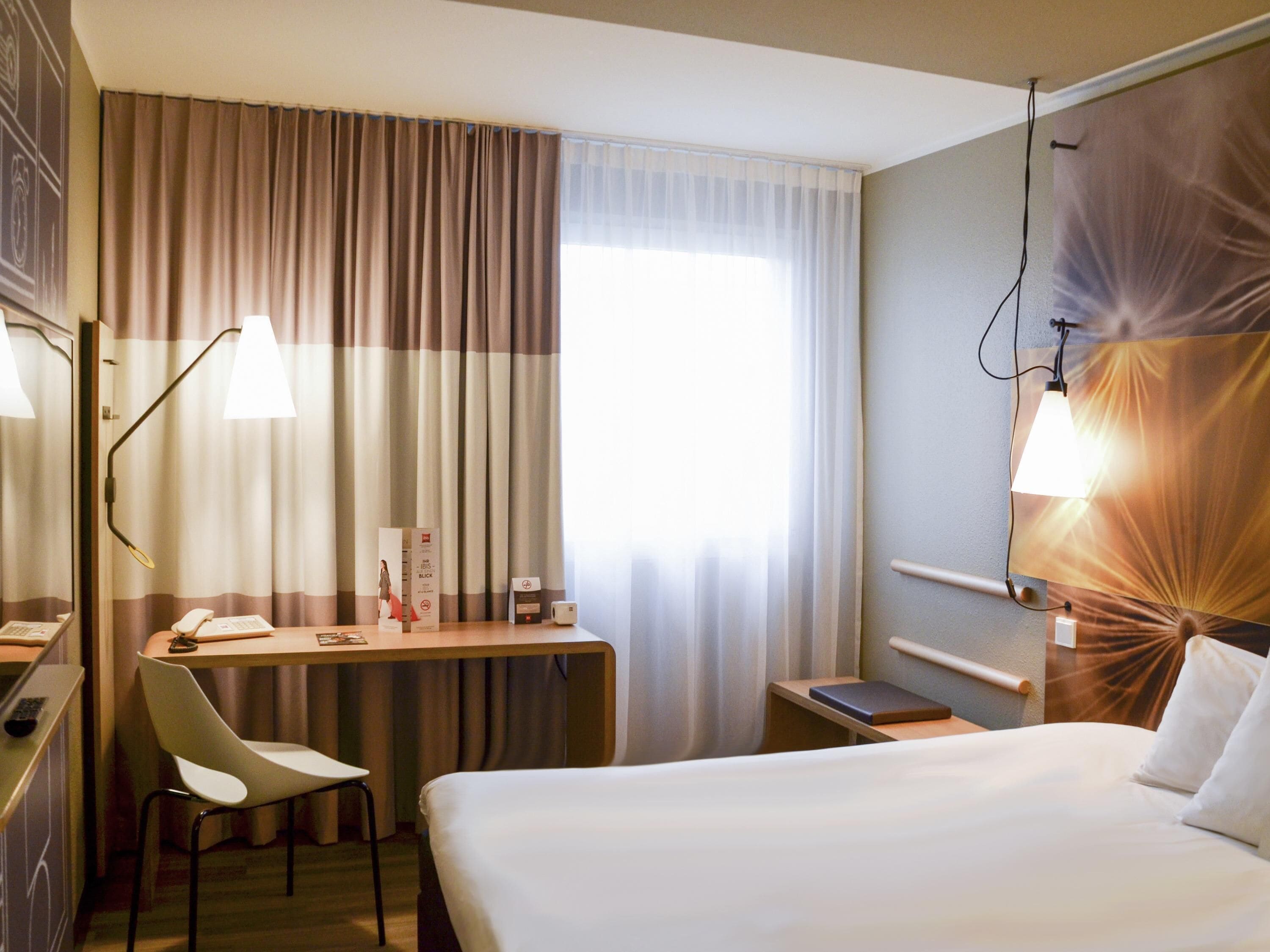 A hotel room with a bed, desk, chair, and a wall-mounted lamp.