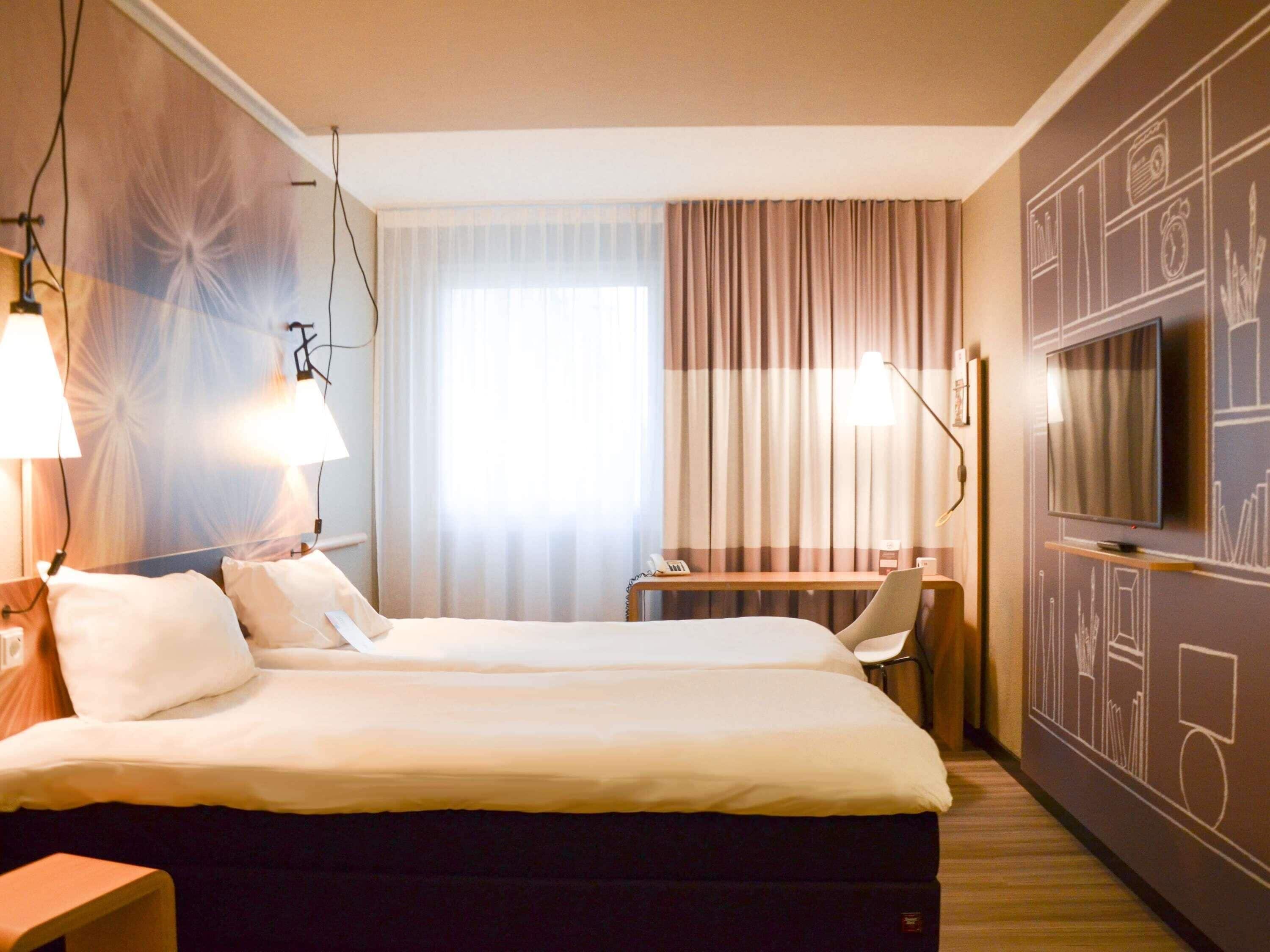 A hotel room with a bed, bedside lamps, a desk, a TV, and a curtain.