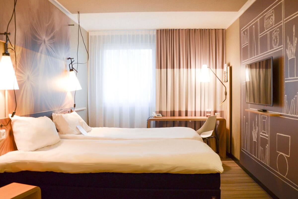 A hotel room with a bed, bedside lamps, a desk, a TV, and a curtain.