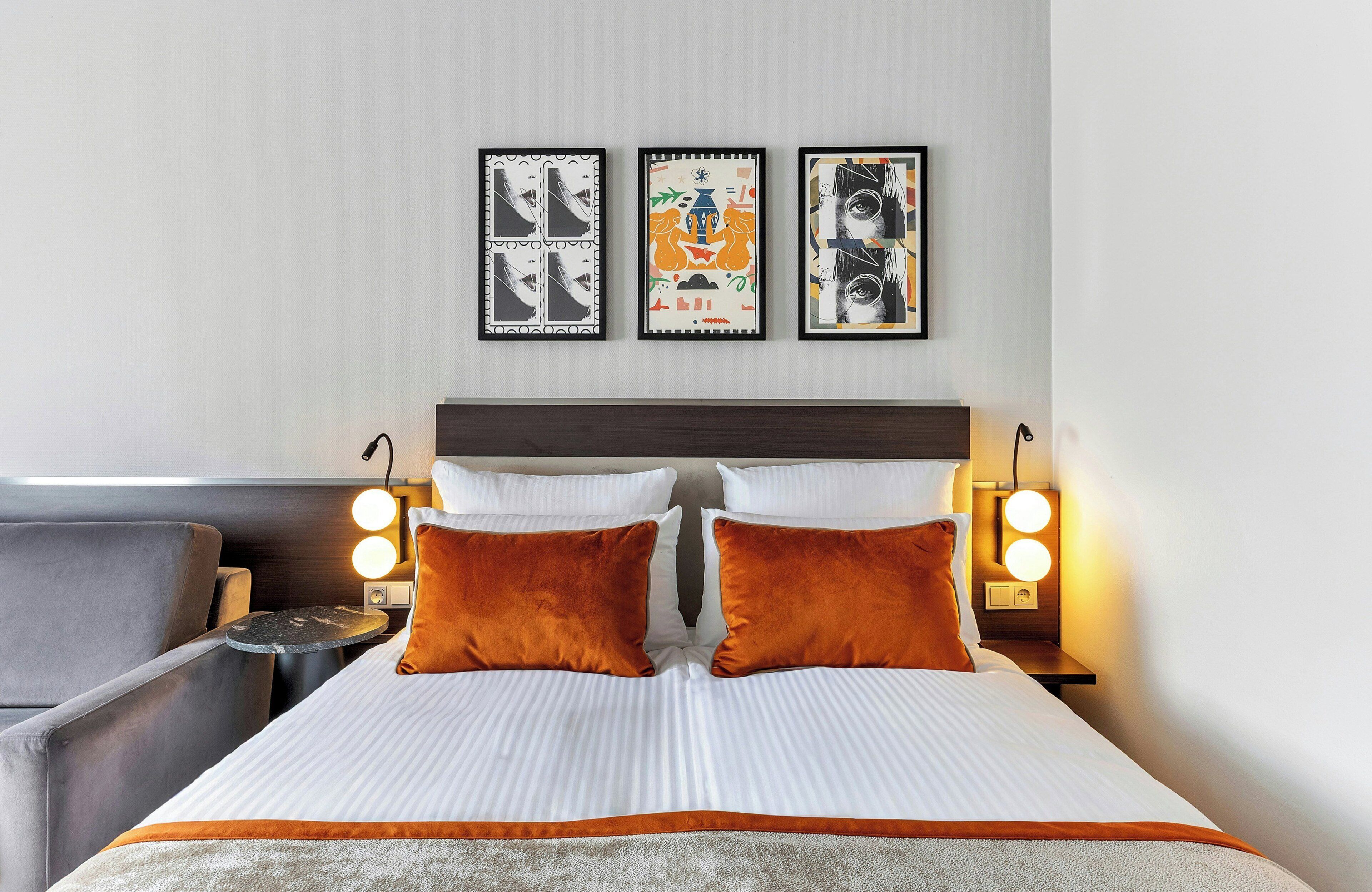Habitación de hotel con cama, dos almohadas, mesita de noche, lámpara, silla y tres cuadros en la pared.