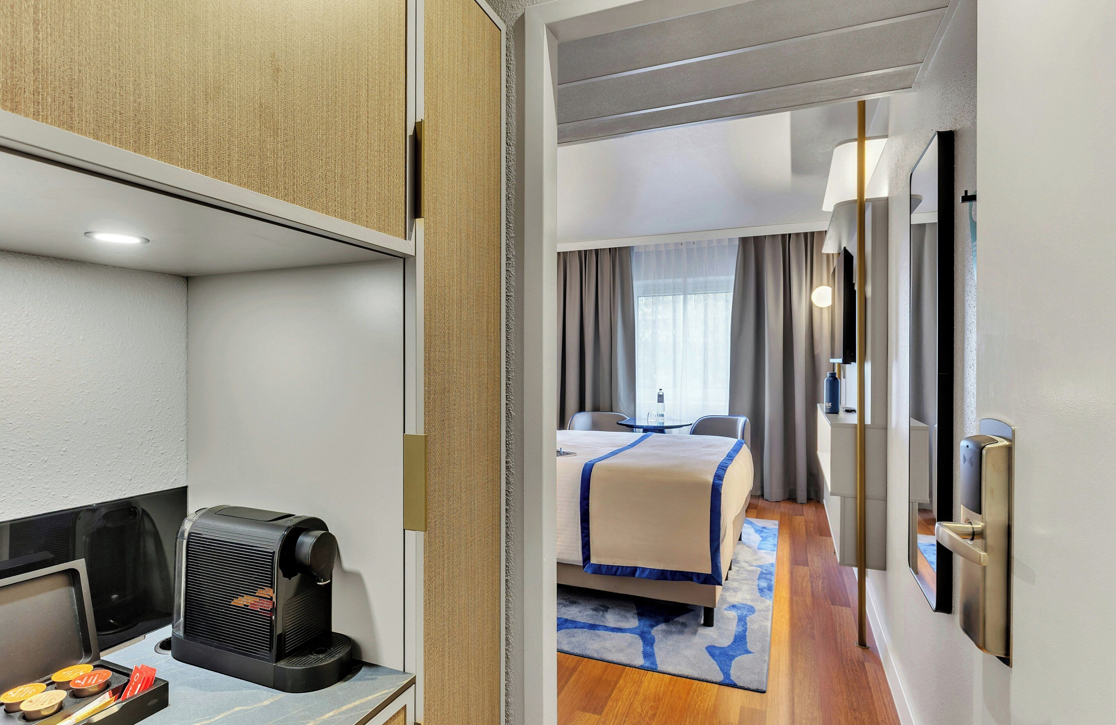 Habitación de hotel con cama, escritorio con cafetera, espejo y puerta con cerradura.