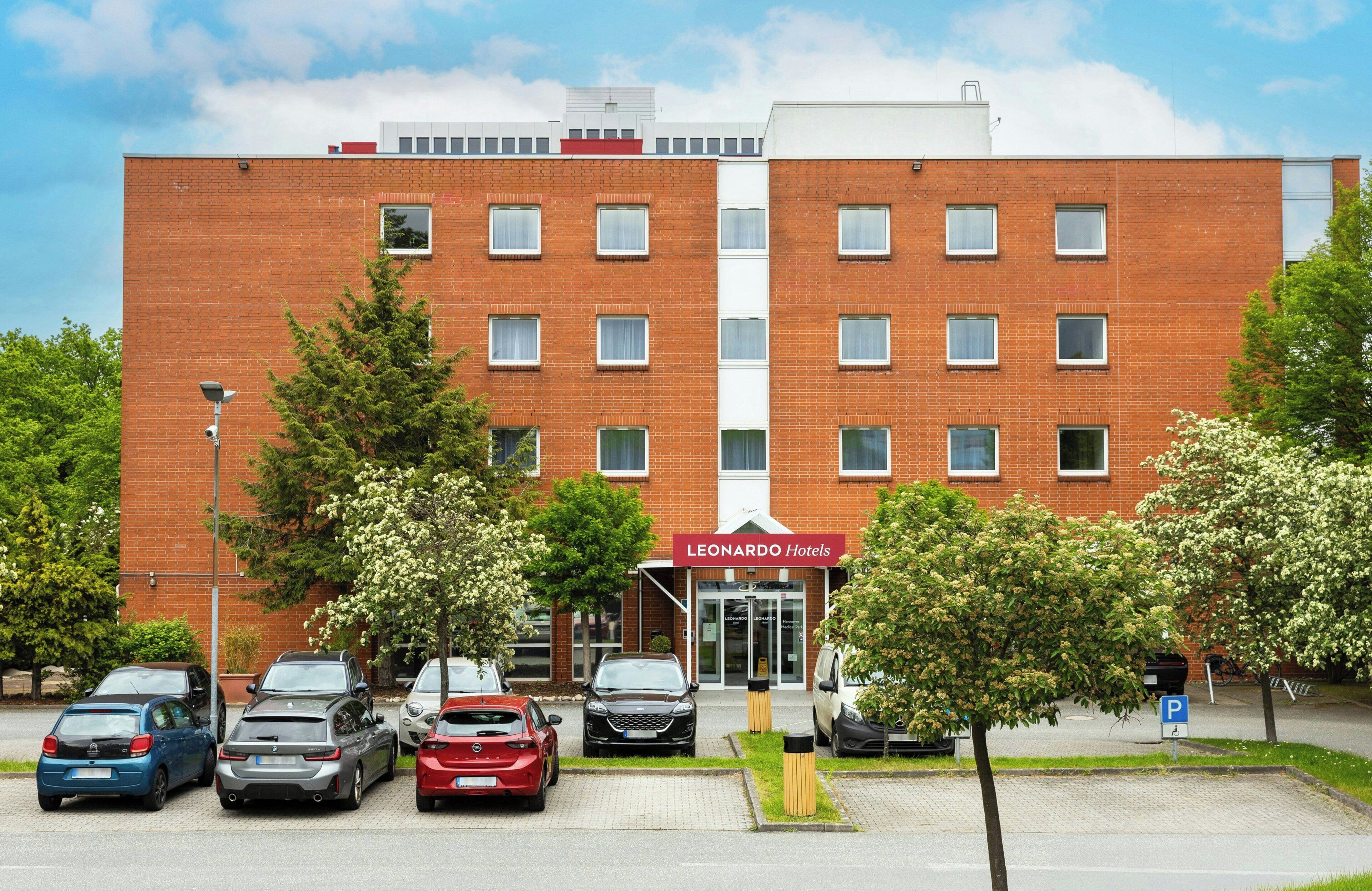Ein Hotel mit roter Backsteinfassade, zahlreichen Fenstern und einem Parkplatz mit mehreren Autos.