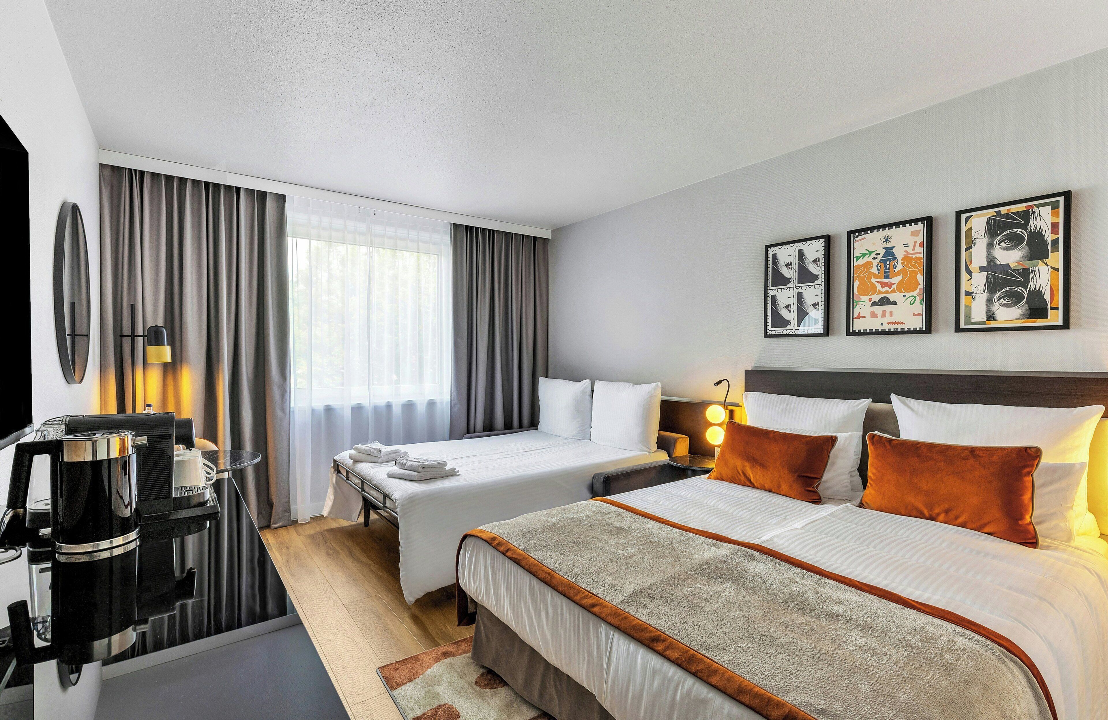 Habitación de hotel con una cama, un escritorio con cafetera, televisor y tres cuadros en la pared.