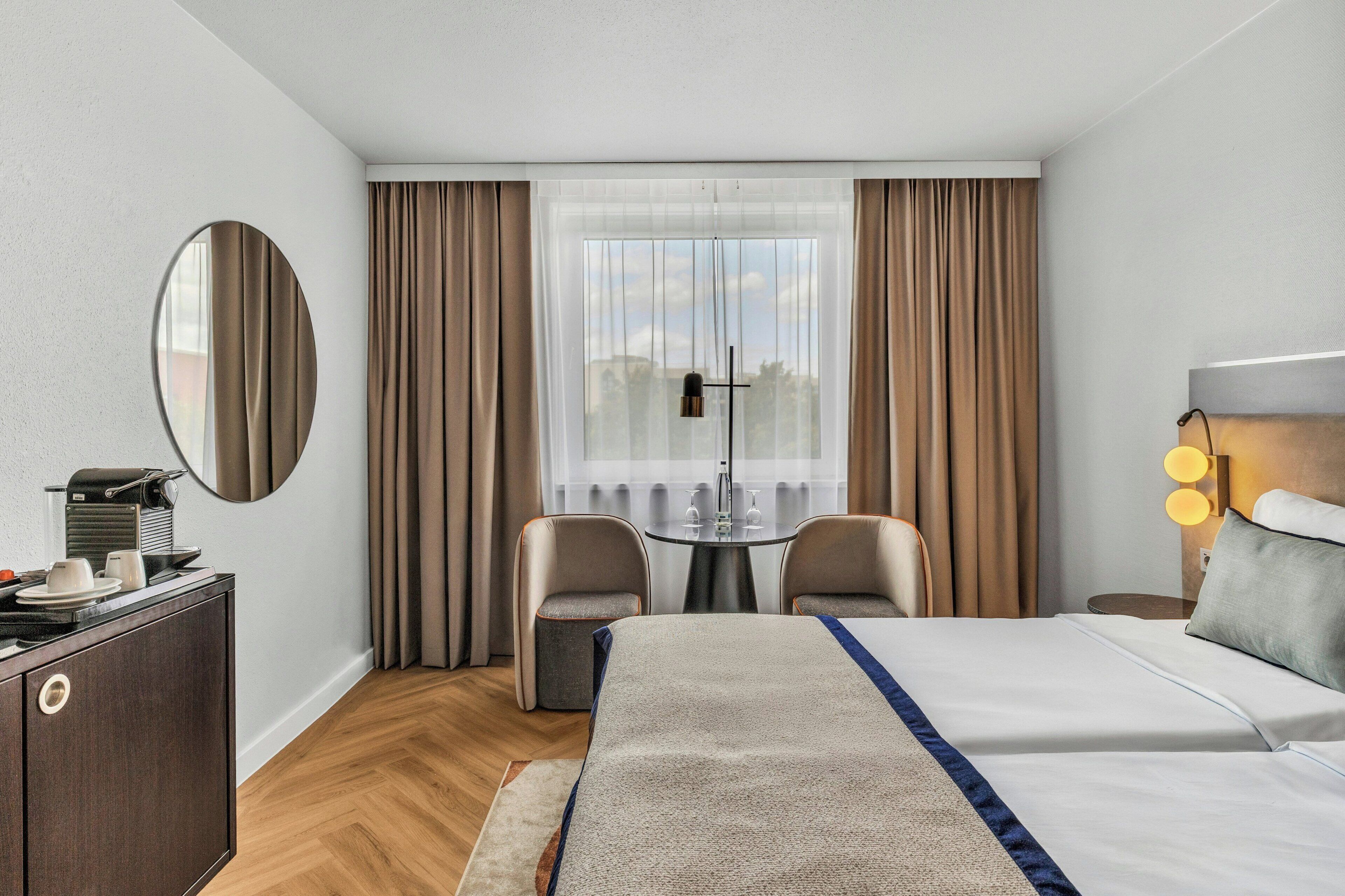 A hotel room with a bed, a desk, a chair, a round mirror, and a window with curtains.