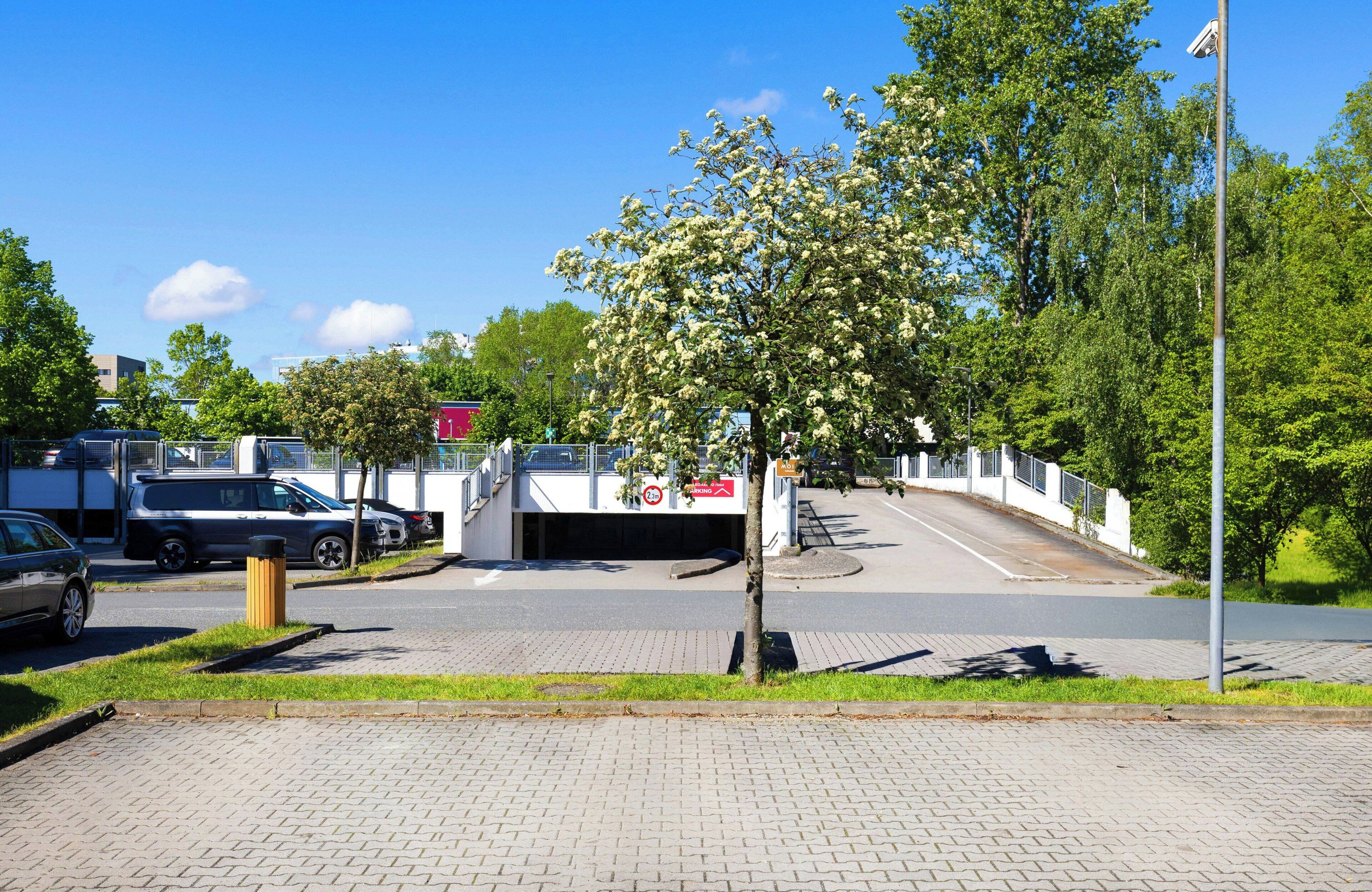 Een parkeerplaats met een boom op de voorgrond, een gebouw met een rood bordje achterin, en een heldere blauwe lucht.
