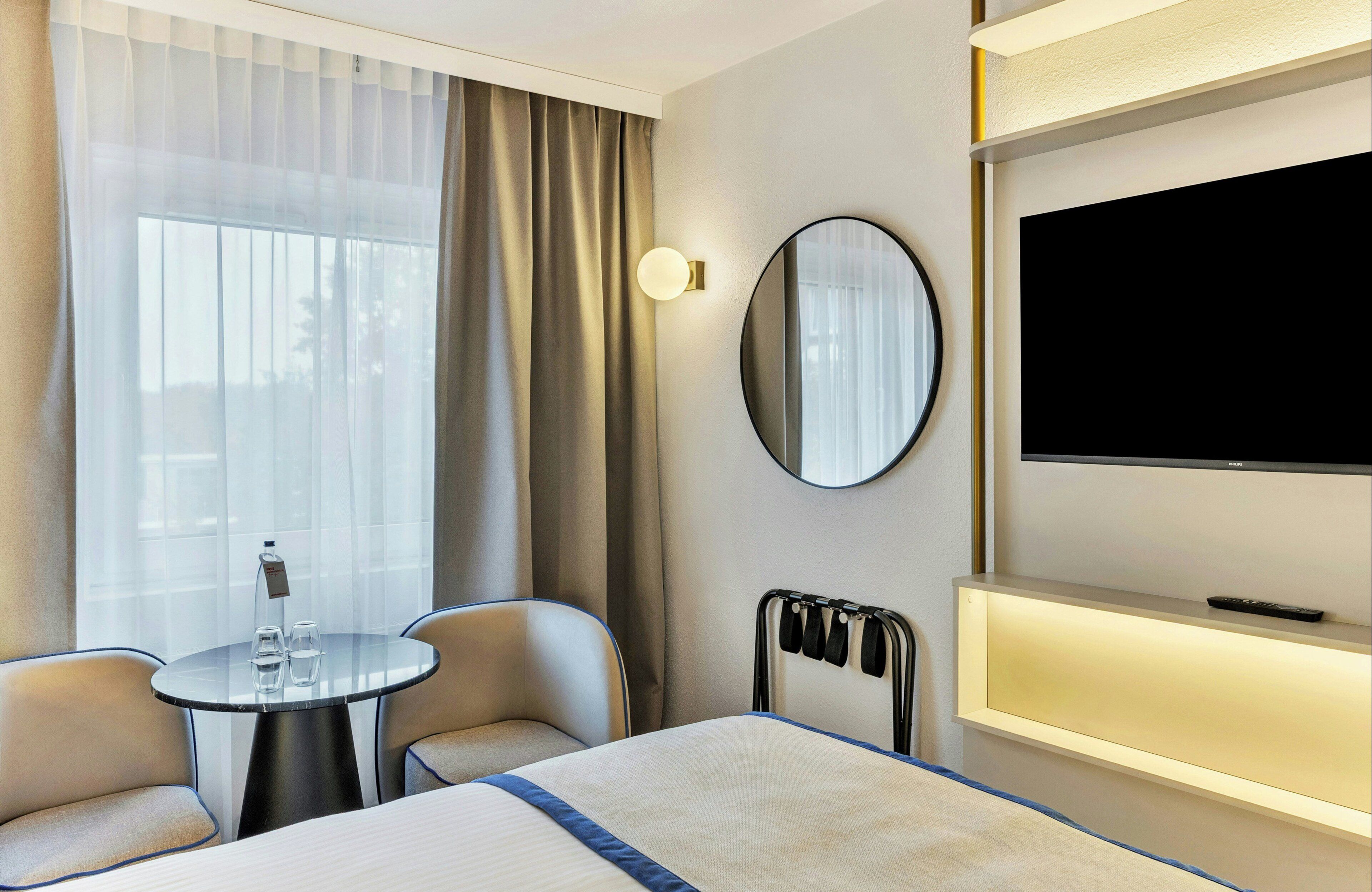 Ein Hotelzimmer mit Bett, Fernseher, Nachttisch mit Gläsern, Stuhl und einem Fenster mit Vorhängen.